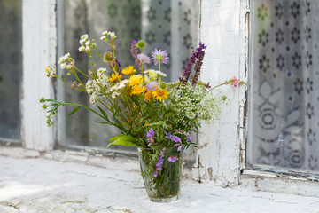 A bouquet of modest wild steppe flowers in an ordinary glass on the windowsill of an old house. Shallow depth of field