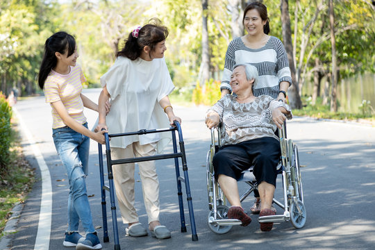 Happy asian family,senior woman, disabled grandmother with walker and wheelchair, daughter,teen granddaughter enjoying a walk in outdoor park, child girl and mother supporting,caring for the elderly