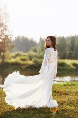 Fototapeta na wymiar Beautiful slender woman in a long white dress walks in the morning near the lake. Brown-haired girl with long hair walks on the grass in the village, natural cosmetics and makeup