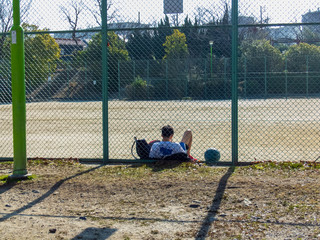 バスケットボールの練習後、グラウンドのフェンスにもたれて休む少年