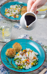 Rice with vegetables and fishcakes on bright plates with glasses of water and lemon, soy sauce in a sauceboat on a gray table background