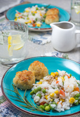 Rice with green peas and corn with fishcakes on bright plates with glasses of water and lemon, soy sauce in a sauceboat on a gray table close-up background