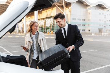 Image of young businesslike man and woman putting luggage in car trunk