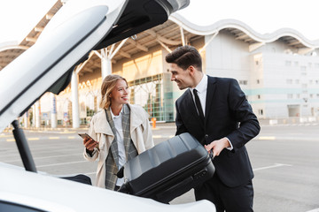 Image of young businesslike man and woman putting luggage in car trunk