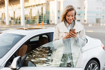Image of businesslike woman standing by car and using cellphone outdoors - obrazy, fototapety, plakaty