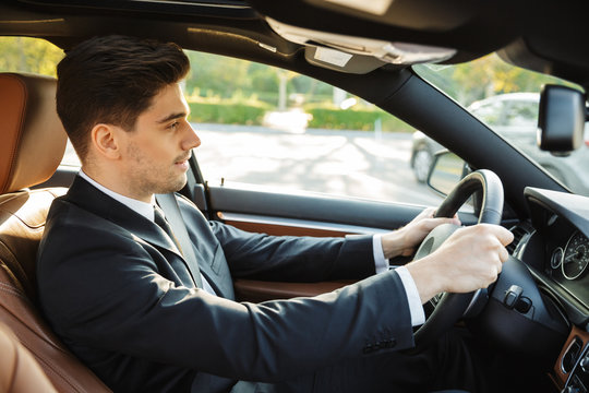 Image of young caucasian businesslike man in black suit driving car