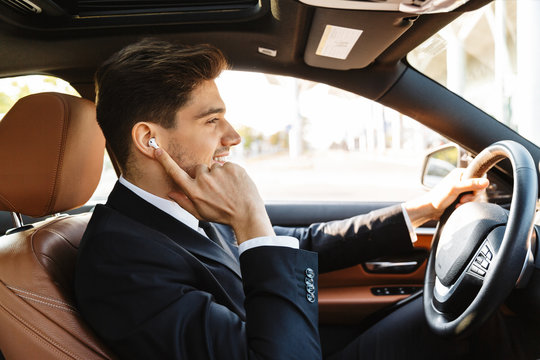 Image of young businesslike man in suit using earbuds while driving car