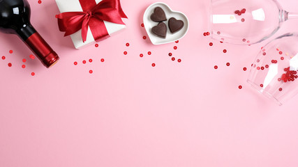Happy Valentine's Day card template. Frame made of bottle of wine champagne, white gift box with red ribbon bow, heart shaped candies, glasses on pink background. Flat lay, top view.