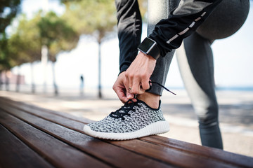 A young sportswoman with smartwatch outdoors in city, tying shoelaces. - Powered by Adobe