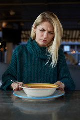 Blonde Caucasian woman in green pullover demonstrates disgust twisting face with negative reaction while trying to eat some smelly soup from a plate on a table