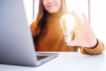 Closeup image of a businesswoman holding and showing a glowing light bulb while working on laptop