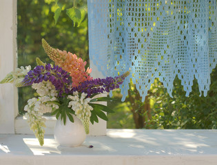 Bouquet of colorful lupines in a white vase on an open window with blue lace curtains, looking out into the summer sunny garden