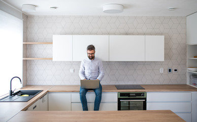 A mature man with laptop sitting in kitchen in unfurnished new house.