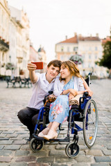 Young woman in wheelchair and her husband taking selfie outdoors, walking in old city center