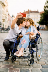 Young Couple taking Selfie. Pretty happy young disabled woman in wheelchair kissing with her young boyfriend and smiling while taking selfie with him In The City
