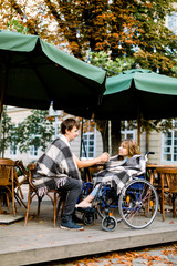 Portrait of pretty young woman on a wheelchair having a good time during a date with her handsome man, drinking coffee and holding hands, sitting in cafe outdoors in the city