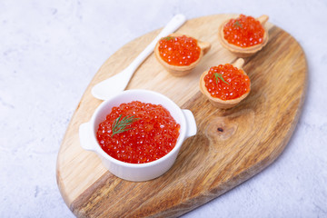 Red caviar (salmon caviar) in a white bowl on a wooden board. In the background are tartlets with caviar. Selective focus, close-up.