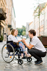 Handsome happy man smiling and holding hand of his beautiful beloved blond handicapped woman in wheelchair while walking together on the street of old city