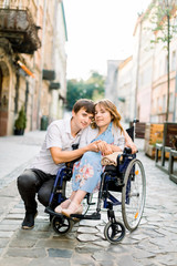 Pretty young smiling woman in the wheelchair and handsome man looking at camera, holding hands, in love, walking in the old city center.
