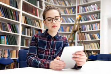 Girl in glasses working with tablet sitting in library. Student prefers to work with a tablet than books