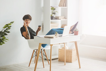 Pretty young businesswoman sitting in a chair with the legs over the desk and talking on phone.