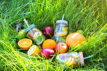 Fresh lemonade in jars with straws. Hipster summer drinks with fruit in green grass outdoors.  Eco-friendly in the nature. Lemons, oranges and berries with mint in the glass. Healthy vegan lifestyle.