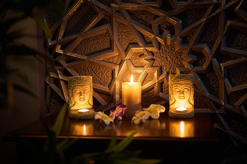 There is a candle on the table, on the sides of the candle are two statuettes of the Buddha, behind the candles is a golden tree. Thai style