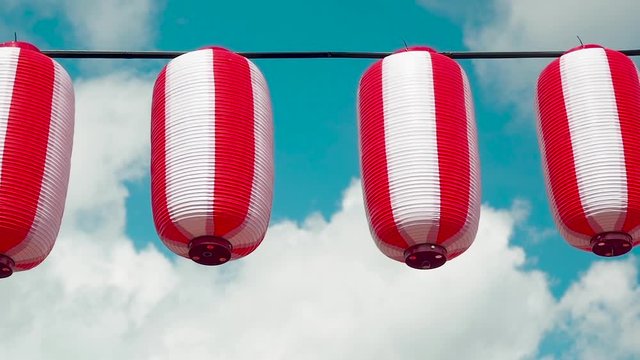 Oriental paper red-white lanterns Chochin hanging on white cloudy blue sky background. Japanese lanterns hanging on bright sky background. Pan right