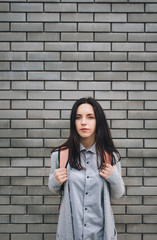 Beautiful young woman with a backpack standing half a turn near the gray brick wall. Lonely look. Pensive girl. Copy space.