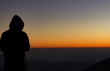 silhouette of Travel photographer standing with a camera mounted on a tripod and shooting a time lapse of the sunrise/sunset. man wearing his hood enjoying the mountain view /valley view