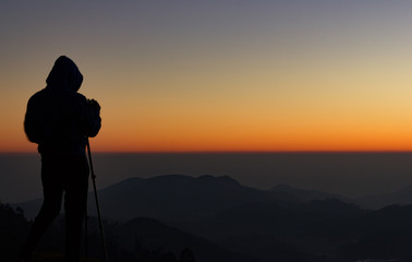 silhouette of Travel photographer standing with a camera mounted on a tripod and shooting a time lapse of the sunrise/sunset. man wearing his hood enjoying the mountain view /valley view