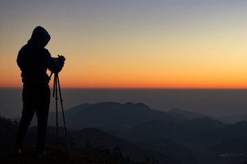 silhouette of Travel photographer standing with a camera mounted on a tripod and shooting a time lapse of the sunrise/sunset. man wearing his hood enjoying the mountain view /valley view