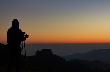 silhouette of Travel photographer standing with a camera mounted on a tripod and shooting a time lapse of the sunrise/sunset. man wearing his hood enjoying the mountain view /valley view.