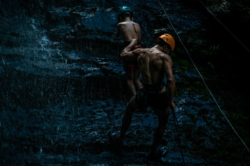 Rappel, hombre descendiendo por montaña con un niño amarrados por una cuerda, mientras cae agua sobre las piedras de noche