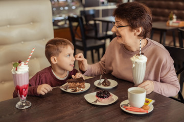 A happy grandmother with glasses and her grandson spend 7 years together in a cafe. they eat sweet cakes and milkshakes.