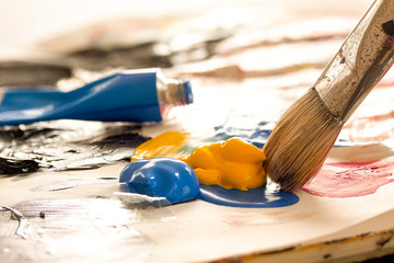 Artist brush with paint from painting tubes ready to mix on a palette. Blue color tube in the background. Backlit close-up image with selective focus.