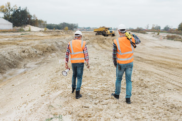 Obraz na płótnie Canvas Two professional road construction workers in orange vests and protective helmets in the middle on the terrain