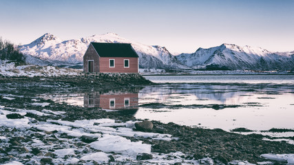 Amazing winter landscape with red rorbu, traditional scandinavian fishing house, snowy mountains in sunset light, blue sky and reflection in water, Lofoten Islands, Norway. Outdoor travel background