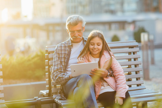 Portrait of a grandfather and granddaughter sitting on a bench and using laptop and smiling.
