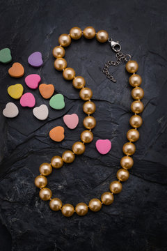A single strand golden color pearl necklace lies on a rough black slate background. For Valentines Day the necklace is accented by small colorfull candy hearts.