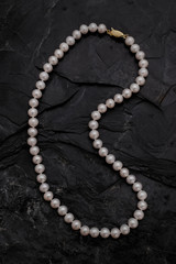 A single strand necklace of round white pearls with a gold clasp curves across a black rough...