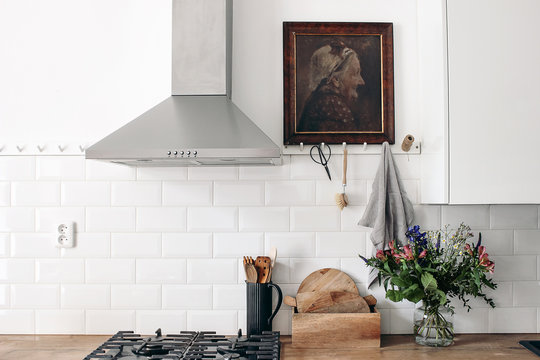 Modern eclectic kitchen interior. White brick wall with metro tiles, peg rails and oil painting. Wooden countertop, stainless steel hood and gas stove. Scandinavian design. Home staging, concept.