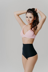 Obraz premium Vertical shot of sexy lady in sexy bra and black pants, has slim perfect figure, dark hair combed in pony tail, models against grey background, looks straightly at camera, has beautiful body