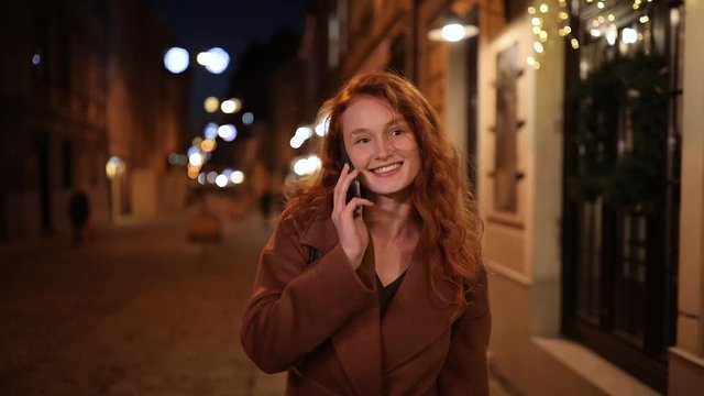 Smiling red-haired girl talking on the phone walking down the street of a night city. Woman smiling and communicating with friends