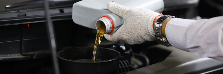 Focus on auto mechanic in gloves replacing and pouring fresh oil into engine at automobile service...