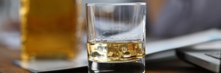 Focus on glassful with alcoholic beverage with ice-cube on wooden brown table. Relax after tough day. Scotch or whisky gourmet alcohol. Blurred background