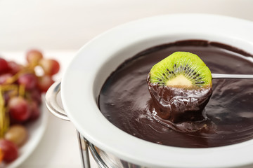Dipping of tasty kiwi into bowl with chocolate fondue on table, closeup