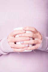Beautiful female hands holding white cup on the pale violet background. Manicure with pink color nail polish with shiny design, nude manicure. Copy space