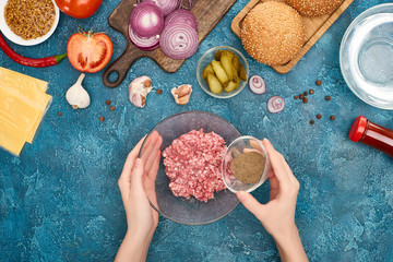 top view of woman adding black pepper to minced meat near burger ingredients on blue textured surface