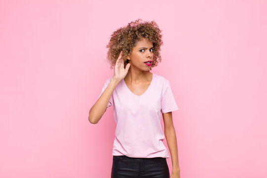 young african american woman looking serious and curious, listening, trying to hear a secret conversation or gossip, eavesdropping against pink wall